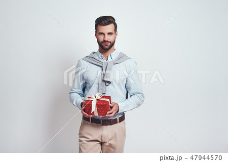 Surprise! Charming bearded man is holding a gift box and smiling while standing in studio on a grey Surprise! Charming bearded man is holding a gift box and smiling while standing in studio on a grey 47944570
