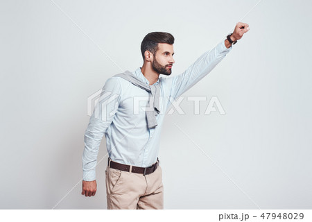 Superhero. Young bearded man is raising his hand up while standing against grey background 47948029