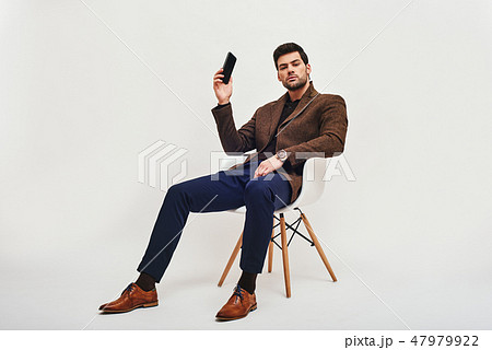 I have an idea! Stylish dark-haired man sitting on a chair, holding a phone in his hand and looking I have an idea! Stylish dark-haired man sitting on a chair, holding a phone in his hand and looking 47979922
