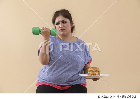 Sad Overweight Woman holding a burger and dumbbell Sad Overweight Woman holding a burger and dumbbell 47982452