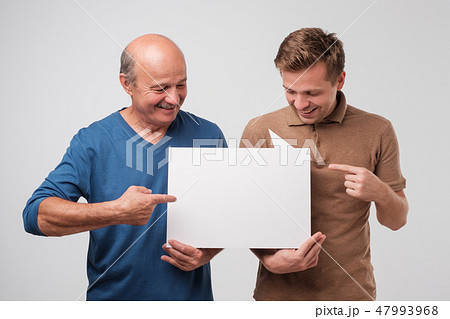 Two mature men father and son are holding a white empty billboard together Two mature men father and son are holding a white empty billboard together 47993968