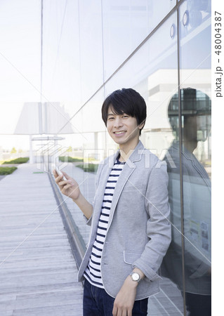 ガラスの壁に寄り掛かってスマホ操作する大学生 48004387
