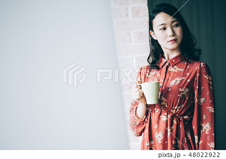 マグカップを手に窓際に立つ赤いワンピースの女性 48022922