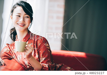 窓際のソファに座る髪をアップにした赤いワンピースの女性 48023227