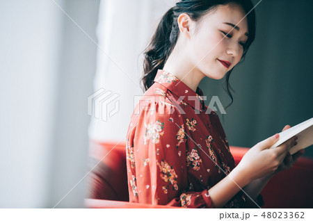窓際のソファに座り本を読む髪をアップにした赤いワンピースの女性の写真素材