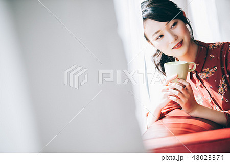 窓際のソファに座る髪をアップにした赤いワンピースの女性 48023374