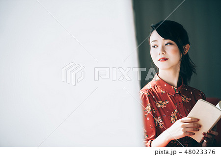 窓際のソファに座り本を読む髪をアップにした赤いワンピースの女性 窓際のソファに座り本を読む髪をアップにした赤いワンピースの女性 48023376