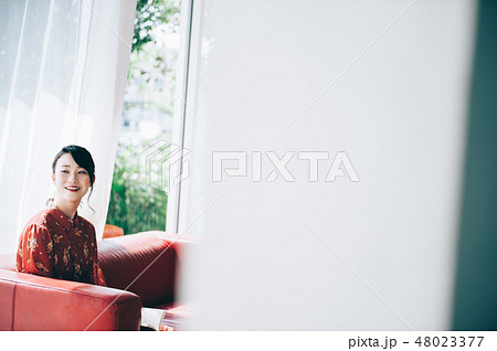 窓際のソファに座り本を読む髪をアップにした赤いワンピースの女性 窓際のソファに座り本を読む髪をアップにした赤いワンピースの女性 48023377