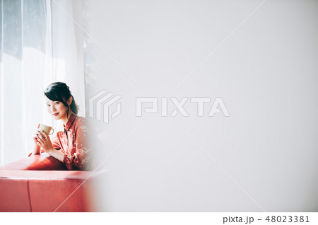 窓際のソファに座る髪をアップにした赤いワンピースの女性 48023381