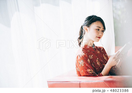 窓際のソファに座り本を読む髪をアップにした赤いワンピースの女性 窓際のソファに座り本を読む髪をアップにした赤いワンピースの女性 48023386