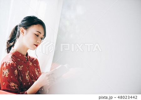 窓際のソファに座り本を読む髪をアップにした赤いワンピースの女性 48023442