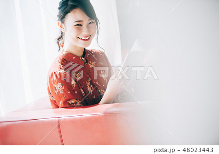 窓際のソファに座り本を読む髪をアップにした赤いワンピースの女性 48023443