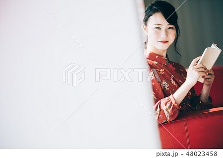 窓際のソファに座り本を読む髪をアップにした赤いワンピースの女性の写真素材