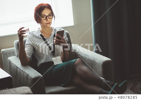 Businesswoman sits in armchair with water glass in hand and talking on phone 48078285
