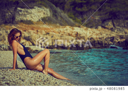 Beautiful tanned girl in a bikini sitting on a rocky beach. Beautiful tanned girl in a bikini sitting on a rocky beach. 48081985