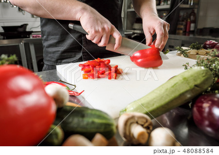 Male chef cuts vegetables for salad in a restaurant in a black apron. White cutting board, closeup Male chef cuts vegetables for salad in a restaurant in a black apron. White cutting board, closeup 48088584