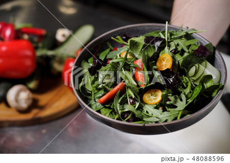 Cook man in a black apron holds a dish of salad in his hand. Against the background of the kitchen Cook man in a black apron holds a dish of salad in his hand. Against the background of the kitchen 48088596