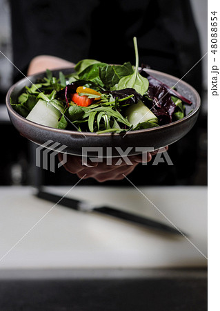 Cook man in a black apron holds a dish of salad in his hand. Against the background of the kitchen 48088654
