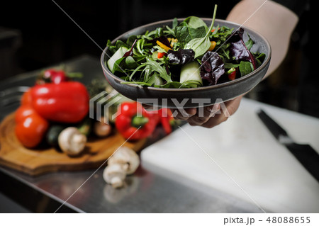 Cook man in a black apron holds a dish of salad in his hand. Against the background of the kitchen Cook man in a black apron holds a dish of salad in his hand. Against the background of the kitchen 48088655