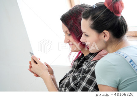 Couple of beautiful young girls in a room with white walls by the window. Communicate and write on 48106826