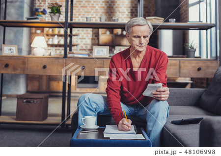 Senior man making notes while sitting in his living room Senior man making notes while sitting in his living room 48106998