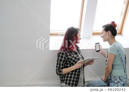 Couple of beautiful young girls in a room with white walls by the window. Communicate and write on 48109242