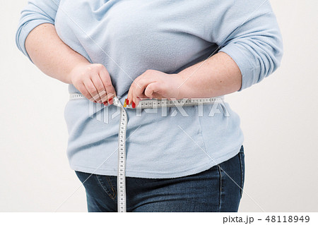 A fat woman measures her waist with a measuring tape in casual clothing on a white background. A fat woman measures her waist with a measuring tape in casual clothing on a white background. 48118949