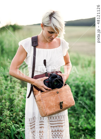 A front view of woman in nature taking out a camera from a brown leather bag. A front view of woman in nature taking out a camera from a brown leather bag. 48121126