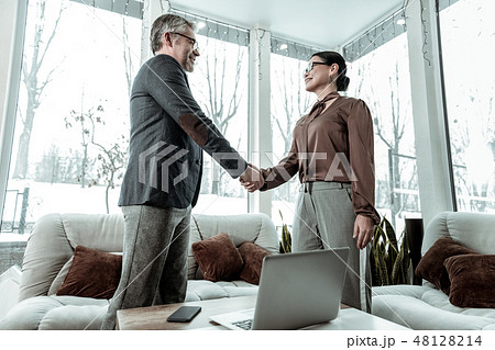 Dark-haired elegant woman in grey trousers and a man in eyeglasses shaking hands Dark-haired elegant woman in grey trousers and a man in eyeglasses shaking hands 48128214