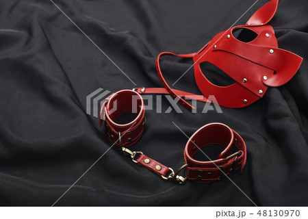 The Pleasures of BDSM. Top view of red bdsm leather set (handcuffs and mask) on a black silk 48130970