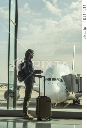 Woman with boarding passes and hand baggage looks out the terminal window on a large airliner 48154155
