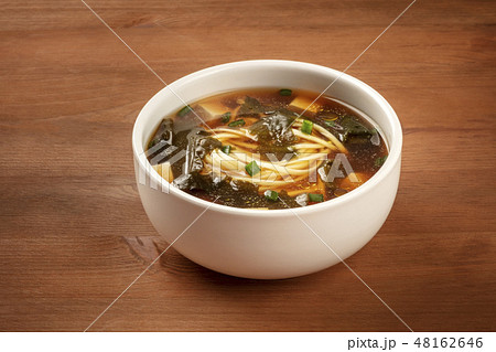 A photo of a bowl of miso soup with tofu, scallions, noodles, and wakame seaweed, on a dark wooden 48162646