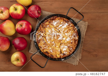 A photo of an apple pie in a pan, shot from the top on a dark rustic wooden background with apples 48162747