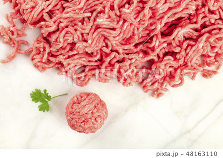An overhead photo of minced meat with a meatball and a parsley leaf, shot from above on a white 48163110