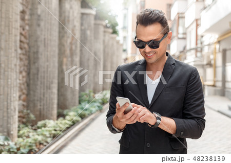 Outdoors leisure. Young man in suit and sunglasses standing on city street using app on smartphone 48238139
