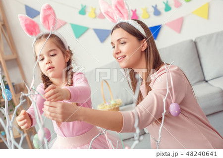 Mother and daughter in bunny ears together at home easter concept hanging eggs on tree Mother and daughter in bunny ears together at home easter concept hanging eggs on tree 48240641