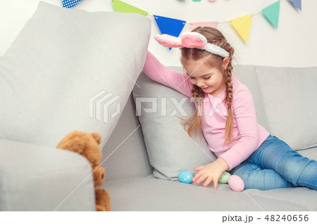Little cute girl in bunny ears at home easter celebration concept hiding eggs under pillow Little cute girl in bunny ears at home easter celebration concept hiding eggs under pillow 48240656
