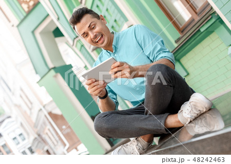 Outdoors leisure. Young man sitting on city street playing game on digital tablet smiling excited Outdoors leisure. Young man sitting on city street playing game on digital tablet smiling excited 48242463