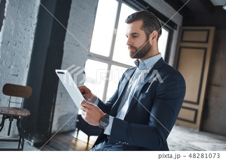 Working hard Confident and young businessman in stylish suit is using his tablet for work Working hard Confident and young businessman in stylish suit is using his tablet for work 48281073