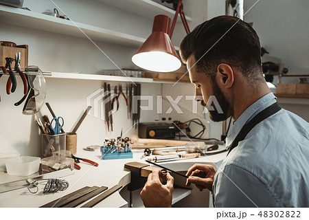 A lot of work in the workshop. Close up of a young male jeweler working and shaping an unfinished A lot of work in the workshop. Close up of a young male jeweler working and shaping an unfinished 48302822