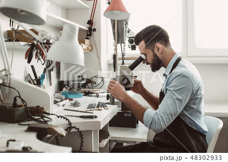 Very close. Side view of a male jeweler looking at the new jewelery product through microscope in a 48302933