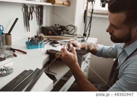 Focused on his work. Close-up photo of male jeweler making a new silver ring at his workbench. 48302947