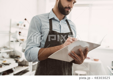Planning a day. Portrait of young male jeweler in apron writing in notebook Planning a day. Portrait of young male jeweler in apron writing in notebook 48303025