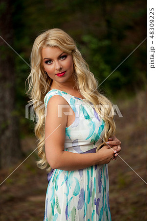Young beautiful blonde woman in white dress with blue and green flowers posing in forest 48309225