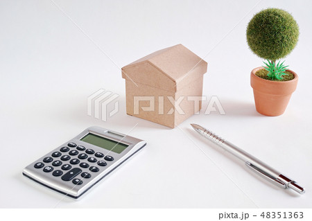 白い紙の上にあるボール紙の家と電卓とボールペンと作り物の丸い鉢植えの木 48351363