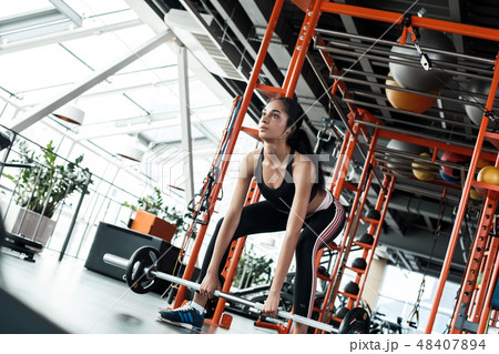 Young woman in gym sporty lifestyle standing holding barbell bending down Young woman in gym sporty lifestyle standing holding barbell bending down 48407894