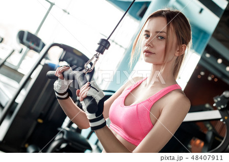 Young girl in sport gloves in gym healthy lifestyle exercising on cable machine holding bar looking Young girl in sport gloves in gym healthy lifestyle exercising on cable machine holding bar looking 48407911
