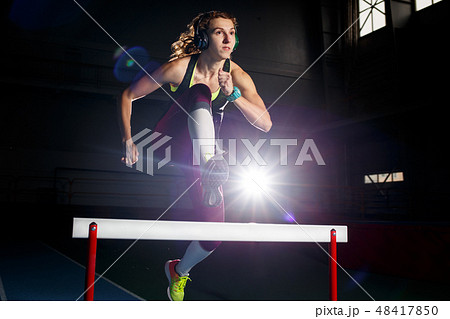 Young sportswoman running and jumping over hurdle 48417850