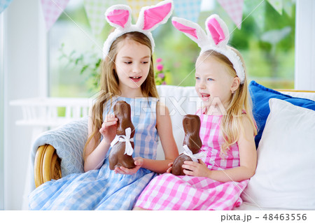 Two cute little sisters wearing bunny ears eating chocolate Easter rabbits 48463556