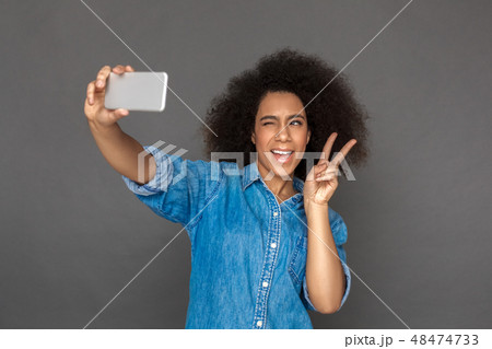 Freestyle. Mulatto woman standing isolated on grey taking selfie on smartphone showing horns sign Freestyle. Mulatto woman standing isolated on grey taking selfie on smartphone showing horns sign 48474733